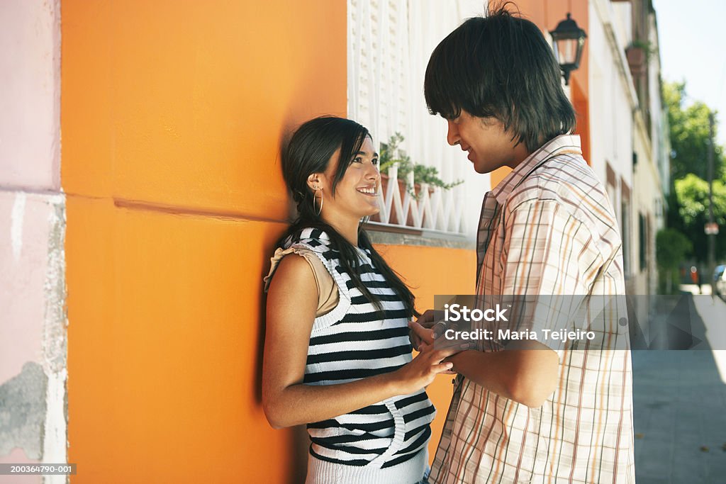 Teenage couple (16-18) holding hands by wall outdoors, smiling  Teenage Couple Stock Photo