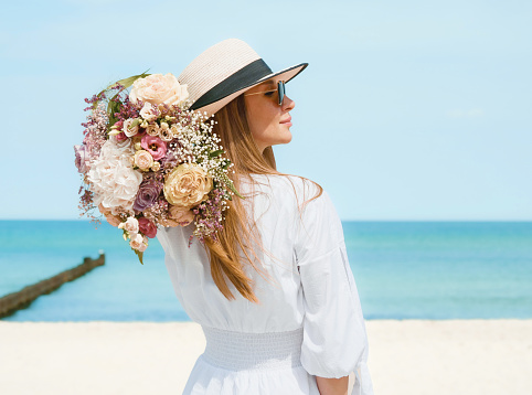 A beautiful young woman stands on a serene beach, the vast expanse of the pristine coastline stretching behind her. She radiates happiness as she holds a vibrant bouquet of fresh, colorful flowers in her hands. The brilliant blue sky forms a striking backdrop, complementing the vivid blooms and enhancing the overall sense of joy and tranquility in this captivating seaside scene. This stock photo captures the essence of relaxation, nature's beauty, and the simple pleasures of life.