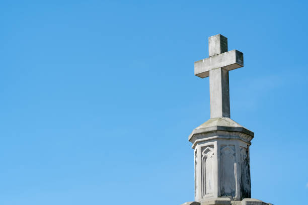 Close-up of a stone religious cross against a blue sky with cloud, three trees, two pines and a cypress, and copy space. Trentino Alto Adige, Italy, Europe.