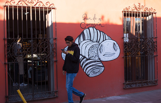 Xochimilco, Oaxaca, Mexico: A young man walks past a mural while looking down at his smart phone; Xochimilco is a neighborhood known for its murals.
