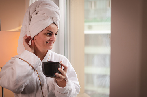 Young businesswoman wearing a robe and a towel looking out the hotel room window while drinking hot coffee.