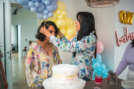 A loving friend places a crown on a smiling Hispanic pregnant woman at her baby shower.