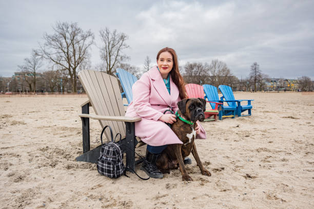 Young lesbian woman with her boxer dog on the beach in winter. She is dressed in casual winter clothing. Exterior of sandy beach in winter on cloudy day.