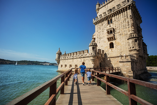 Group of kids travelers excited to visit famous tower of Belem, run small wood bridge toward the entry in castle on sunny day in Portugal