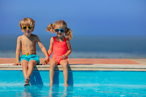 Young friends, boy and girl wear sunglasses sitting comfortably at the brink of a pool, with a serene ocean view in the distance