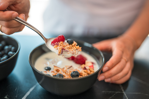 Close up of a full spoon above a black bowl with oatmeal cereal in organic plain cream  and various berries, standing on a marble kitchen counter, another bowl with only blueberries in, next to the first one. The other hand holding the bowl  from the side. High angle shot.