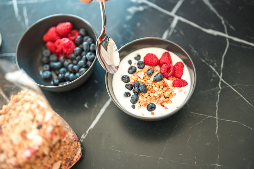Close up of an oatmeal  cereal, yogurt  and berries in a black ceramic bowl, another bowl with only raspberries and blueberries in, next to the first one. A hand holding a small spoon ready to dig in the bowl. High angle shot.