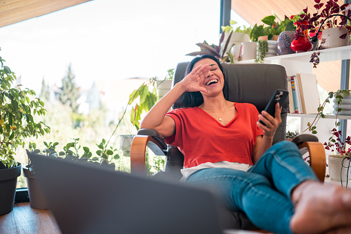 A modern apartment serves as the backdrop for a mid-aged Hispanic woman's remote work, where she effortlessly handles her laptop and smartphone, dressed in casual attire, and exuding a pleasant appearance.
