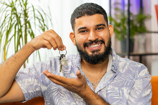 Happy Indian man real estate agent lifting hand showing the keys of new home house apartment, buying or renting property, mortgage loan, investment at modern home room apartment. Guy sits on couch