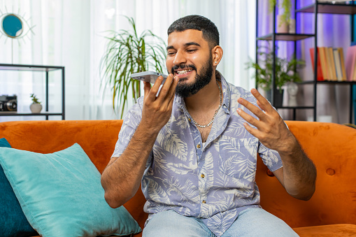 Happy excited Indian man making phone call conversation with friends at modern home room apartment. Arabian guy enjoying mobile loudspeaker talking gossip, rumors, good news sitting on couch indoors