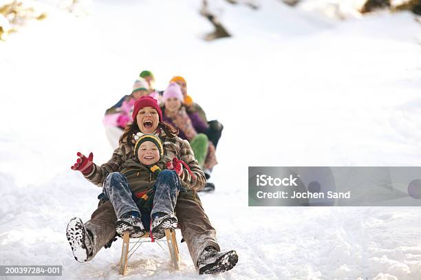 Mother And Son Tobogganing In Snow Family In Background Stock Photo - Download Image Now