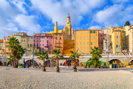 View from Plage des Sablettes beach and promenade of the colorful old town of Menton France, showing the cathedral, bell tower and steps to the basilica and chapel. Menton is a town on the French Riviera in southeast France. It’s known for beaches and gardens such as the Serre de la Madone garden, showcasing rare plants. East, the hilly, medieval old town is home to Basilique Saint-Michel, with its 18th-century bell tower, and the ornate facade of La Chapelle des Pénitents-Blancs.