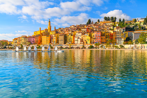 Townscape of the picturesque and colorful old town and Les Sablettes Beach and promenade, with small sailboats sailing in a line along the Cote d'Azur French Riviera at Menton, France. It’s known for beaches and gardens such as the Serre de la Madone garden, showcasing rare plants. East, the hilly, medieval old town is home to Basilique Saint-Michel, with its 18th-century bell tower, and the ornate facade of La Chapelle des Pénitents-Blancs.