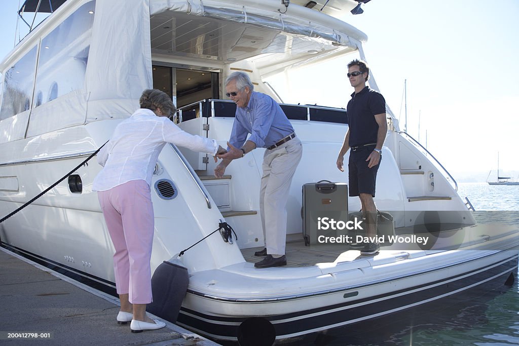 Senior man helping senior woman onto yacht, crew member in background  40-44 Years Stock Photo