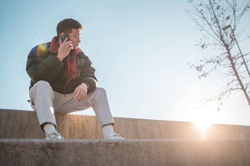 An Asian male teenager in casual attire is engaged in a conversation on his smartphone while sitting on concrete steps outdoors, with a clear sky and the sun low on the horizon.