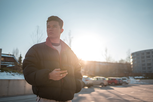 A young Asian male teenager stands outdoors in a winter urban landscape, using a smartphone with a focused expression. He is dressed in casual winter attire with the sun setting in the background.