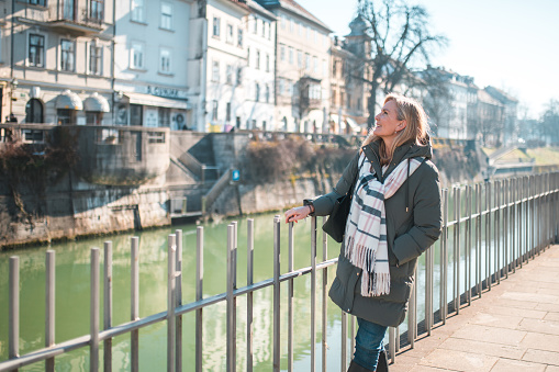 A mature Caucasian woman enjoys sightseeing alone on a sunny day in an urban environment, leaning on a railing with a tranquil river and city architecture in the background. She is dressed in a casual coat and scarf.