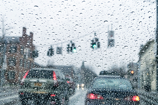 Looking through windshield at cars waiting in traffic just after the stoplight has turned green at a busy road intersection during a cold February winter heavy rain storm.