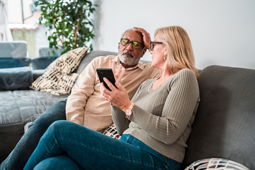 A senior couple is engaged with technology in a cozy living room environment. The mixed race male and his Caucasian female partner are sitting comfortably on a gray sofa, both wearing casual attire, as they focus on a smartphone together.