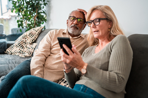 A senior mixed race male and a Caucasian female are seated closely on a couch, intently looking at a smartphone together in a cozy, well-lit living room.
