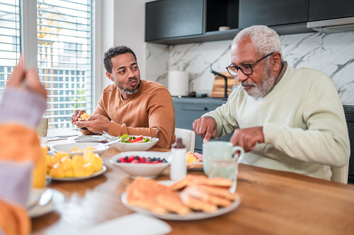 Two men of different generations, likely father and son, sharing a meal during a family brunch in a modern kitchen, representing a diverse multigenerational household.