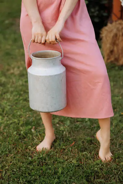 A young adult, caucasian woman in a soft pink dress, holding an old-fashioned metal milk can. Barefoot, standing on a grassy.Concepts dairy products, vintage aesthetics, or rural lifestyle editorials.
