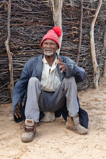 old smiling african man in the yard having a rest, in front of a traditional kraal fence, old casual clothing and a red beanie