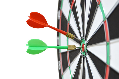 Cutout of two darts, one red and one green hitting bull's eye spot on on a dart board isolated over white background denoting success, win, achievement of business target, competition