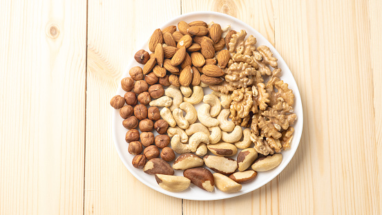 White plate with nuts on wooden background, walnut kernels, peeled dried hazelnuts, almonds and cashews, top view, healthy and wholesome food concept.