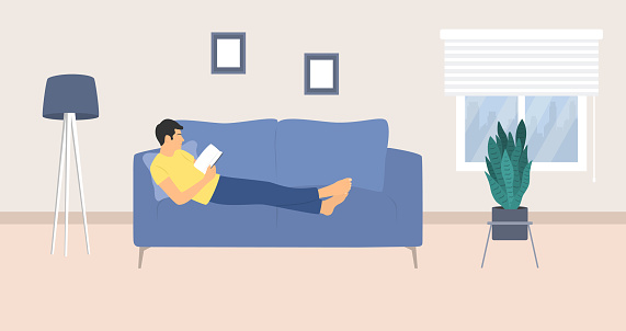 Side View Of Relaxed Young Man Reading Book On Couch. Living Room Interior With Floor Lamp, Sofa And Potted Plant