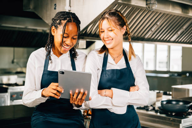 istock Two famous chefs in stainless steel kitchen discuss video blog using tablet. Professionals smiling standing reading searching. Modern technology in restaurant industry holding device. 2004403897