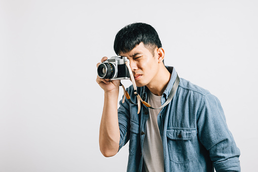 A charismatic young man enjoys a fun photography session with a vintage camera. Studio shot isolated on white background. His captivating look is pure paparazzi style