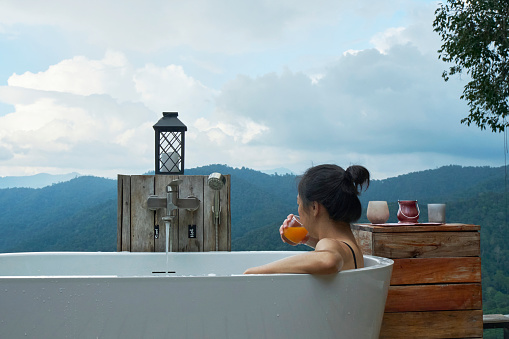 One Asian woman leisurely relaxes in a bathtub and drinks orange juice, resting in panoramic mountain range view of tropical forest, the tranquil unseen scenery natural travel resort for vacation.