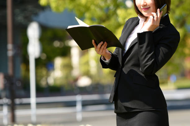 istock Young Asian female office worker in a suit talking on the phone while holding a notebook outdoors 2004433572