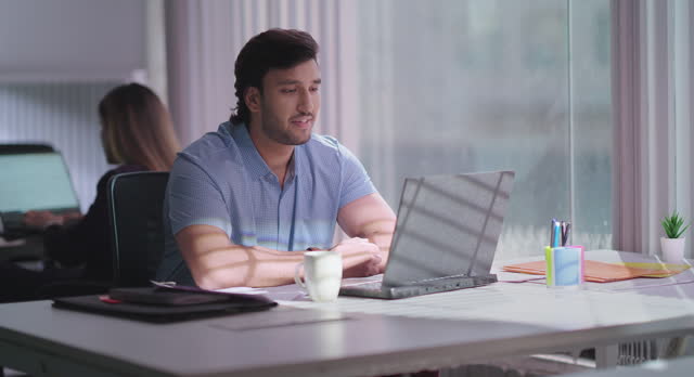 Indian happy busy man wears headphone sitting on chair use laptop look cam talking online video call centre indoor office. Focused male agent provide customer support service communication client at workplace