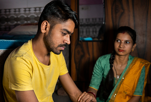 Indoor image of a sad, depressed married Indian young man thinking deeply and his caring wife consoling him at home.