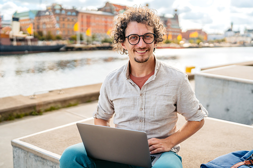 Portrait of a young man sitting on the quayside in Malmo in Sweden and working on his laptop.