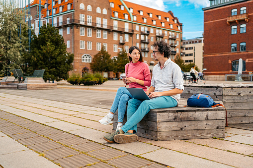 Young male and female students sitting on the quayside in Malmo in Sweden and discussing their class notes on the laptop.