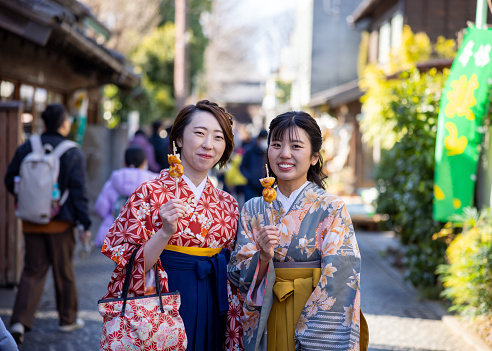 Female friends in Kimono / Hakama eating Japanese ‘Kushi-Dango’ skewered dumplings on street