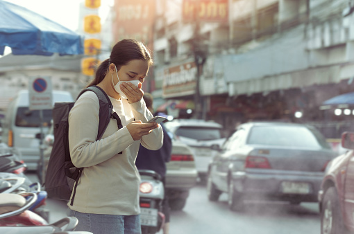 Asian woman wearing PM2.5 dust mask and using phone on the street. wearing mask protect against pollution, anti smog and viruses,  air pollution, and headache suffocating. City air pollution.mist city