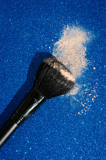 High angle view of a mineral powder with a make-up brush on a shimmering blue background.