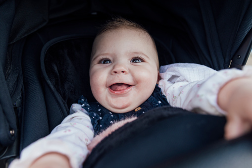 Close up view of a baby girl in a pram, she is looking playful and excited while lying on her back, she is reaching her hands up and has a big smile on her face.