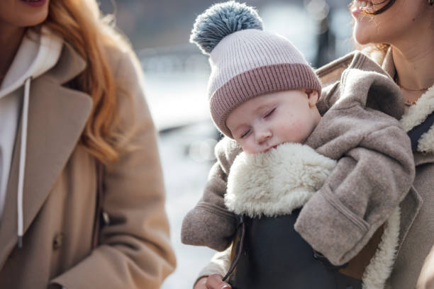 Close up midsection view of two young women walking together along a path with their babies in the city in Newcastle-upon-Tyne, North East England. They are wrapped up in warm clothing on a cold day, One mother has her baby in a baby harness and the baby is sleeping.