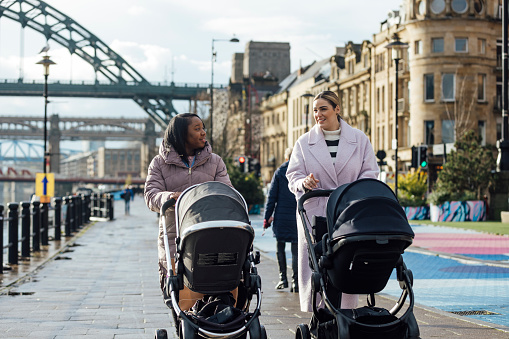 Wide full length view of two young women walking together along a path with their babies in the city in Newcastle-upon-Tyne, North East England. They are wrapped up in warm clothing on a cold day, both mothers have their babies in prams.