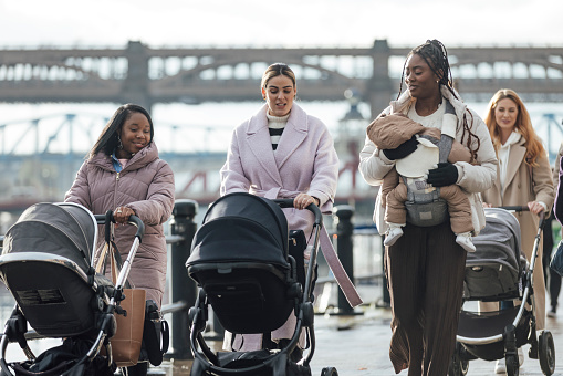 Wide view of a medium group of young women walking together along a path with their babies in the city in Newcastle-upon-Tyne, North East England. They are wrapped up in warm clothing on a cold day, some mothers have their babies in a pram and some have their babies in baby harnesses.