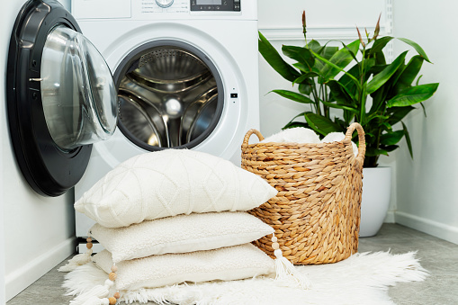 Laundry basket and pile of white pillows in front of the opened washing machine in laundry room. Housekeeping and cleanliness concept