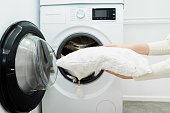 Woman holding clean white pillow in front of the drum of washing machine in laundry room