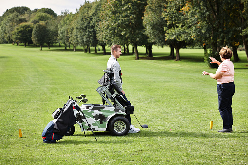 Sports, person with disability and trainer for golf in game, match and competition on golfing course. Recreation, mobility scooter and woman and man with club for training, fitness and field practice