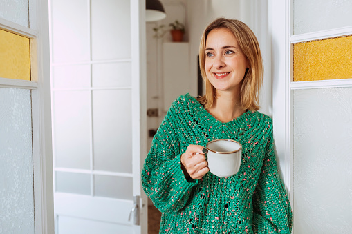 Young beautiful woman with cup of coffee, wears a green sweater, looks away at home. Morning, breakfast concept.