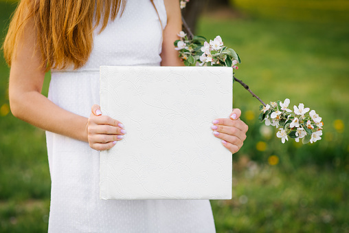 Photo book in a white leather cover in the hands of a woman in spring near a flowering branch of an apple tree